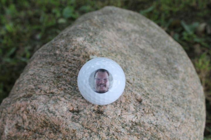 May include: A white golf ball with a picture of a person's face in the center.