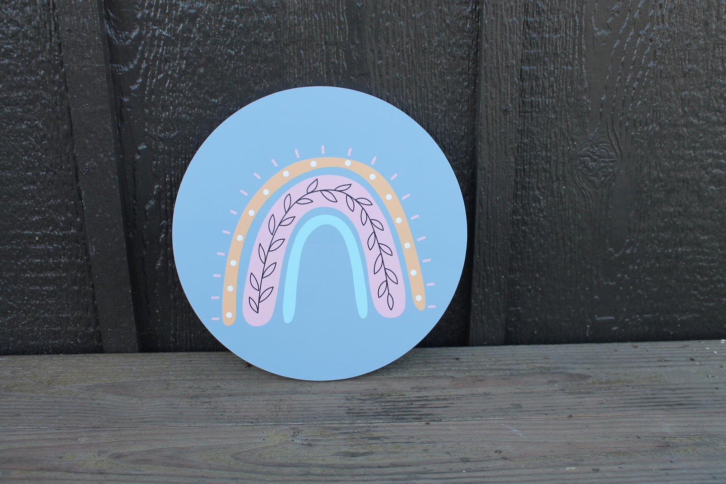 May include: A round wooden sign with a light blue background featuring a rainbow with pink, light blue, and brown accents. The rainbow has a leafy design in the center.