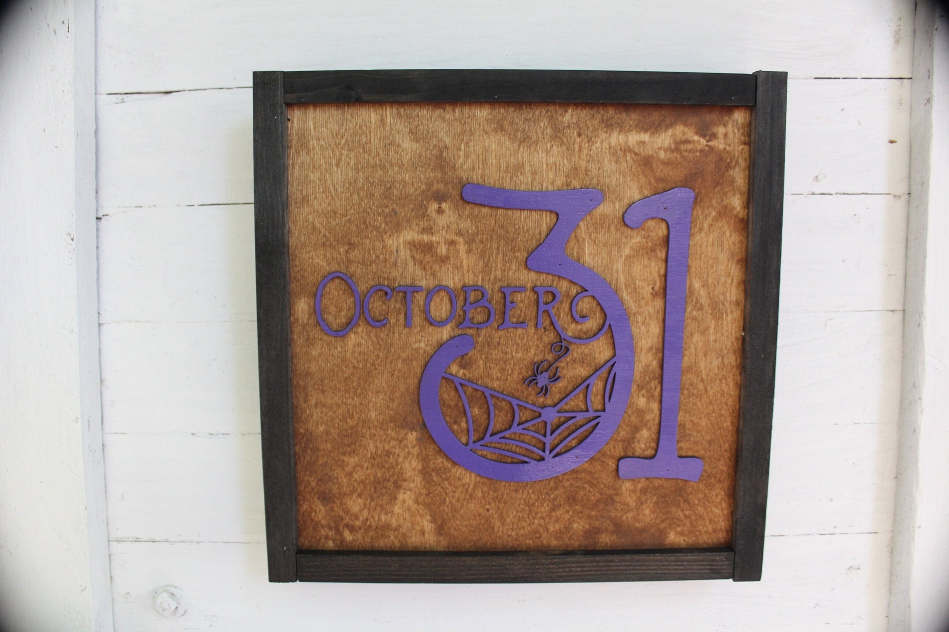 May include: A wooden sign with a black frame and a purple inscription that reads "October 31". The number 31 is stylized with a spider web and spider in the center of the 3.