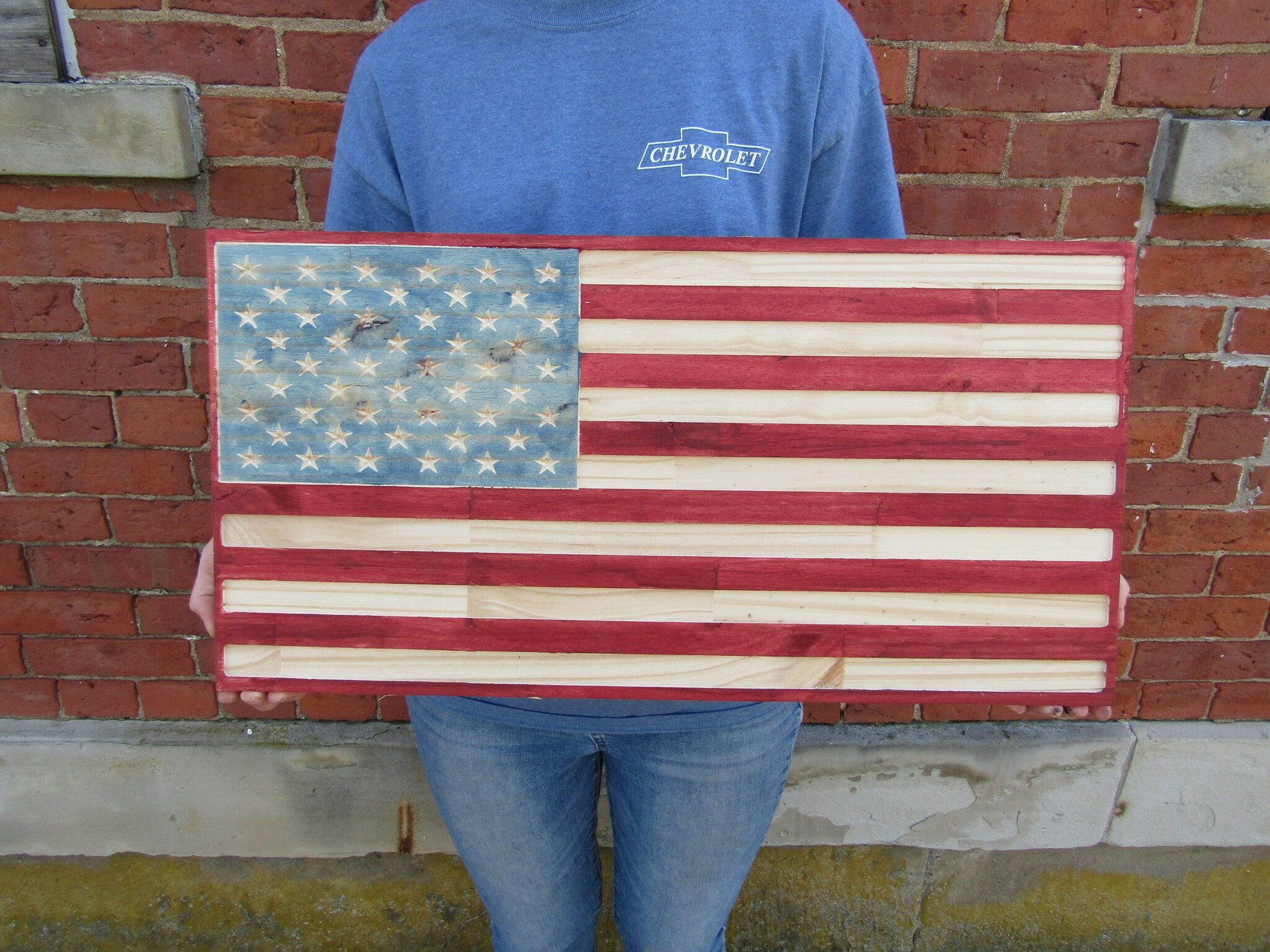 May include: A wooden American flag with red, white, and blue paint. The flag is held in front of a brick wall.
