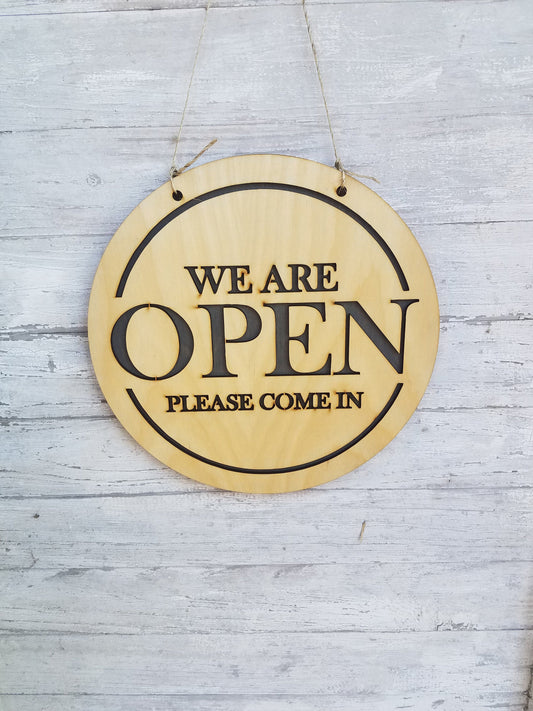 May include: A round wooden sign with the text "We are open. Please come in." in black lettering.